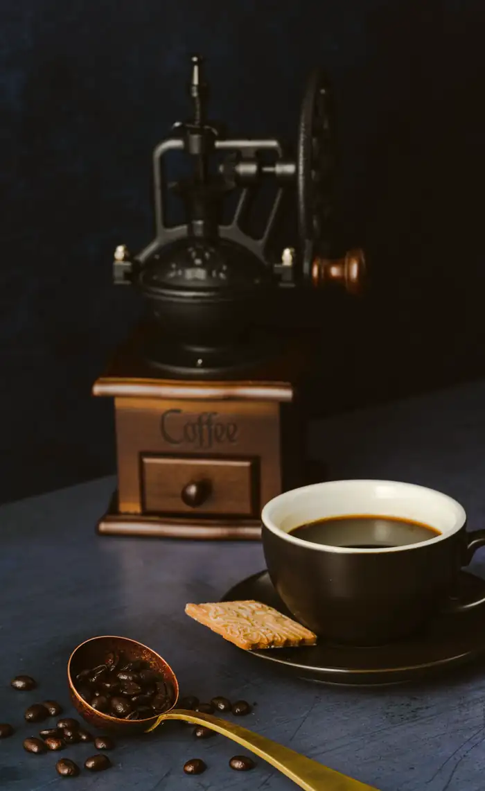 The mill on the bench: a cast-iron grinder with a brass hopper and a hand wheel, a full cup beside it and a scoop of beans on the wood.